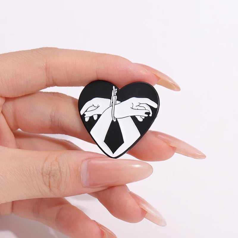 A hand with long, nude-colored nails holds the Kiss Me Later Bound Hands enamel pin—a black and white heart-shaped pin showing two hands bound by rope against a bold black background.
