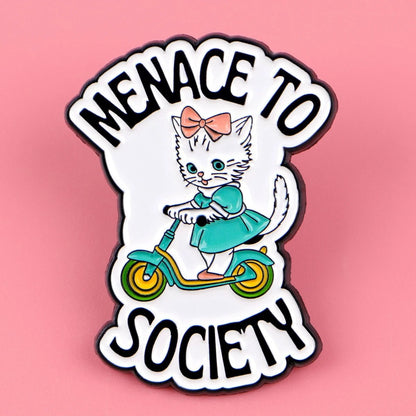 cat enamel pin shows a cute white kitten in a teal dress and pink bow riding a green scooter, with “MENACE TO SOCIETY” in bold black text on a pink background.