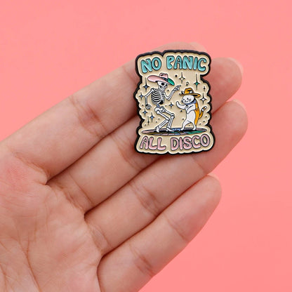 A hand holds a Kiss Me Later enamel pin with skeletons dancing under stars and NO PANIC ALL DISCO text, set on a pink background for major spooky vibes.