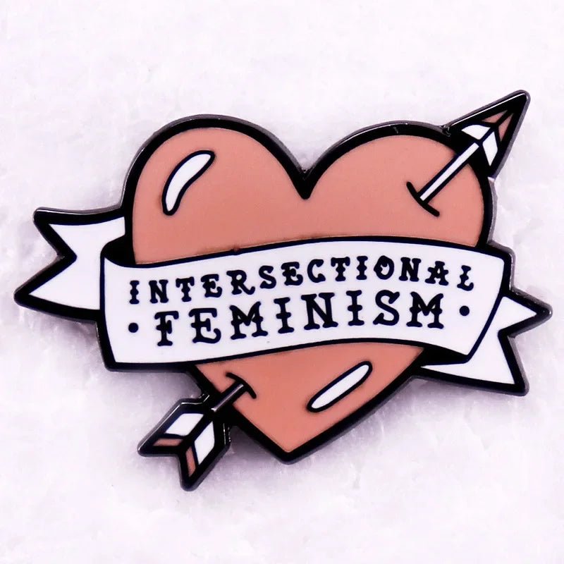 Red pink love heart enamel pin is peach-shaped, pierced by an arrow, and features a white banner with INTERSECTIONAL FEMINISM printed in black letters.