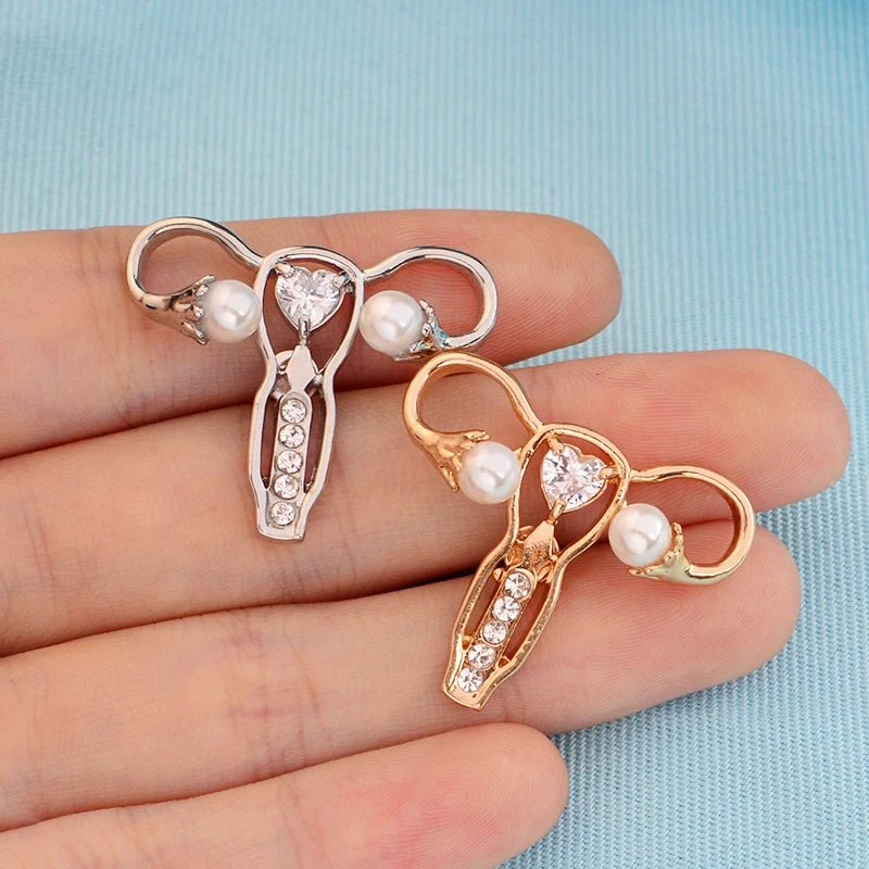 Two uterus ovary pins—one silver, one gold—with pearl accents and rhinestones are shown on a hand against light blue fabric, making a unique feminist gift or medical symbol accessory.