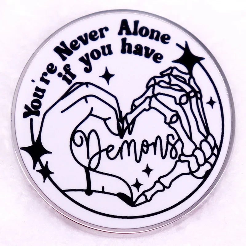 Round pink enamel pin shows a human and skeleton hand making a heart with Demons inside. Above, it reads Youre never alone if you have demons, accented by small stars.