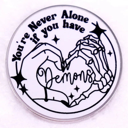 Round pink enamel pin shows a human and skeleton hand making a heart with Demons inside. Above, it reads Youre never alone if you have demons, accented by small stars.