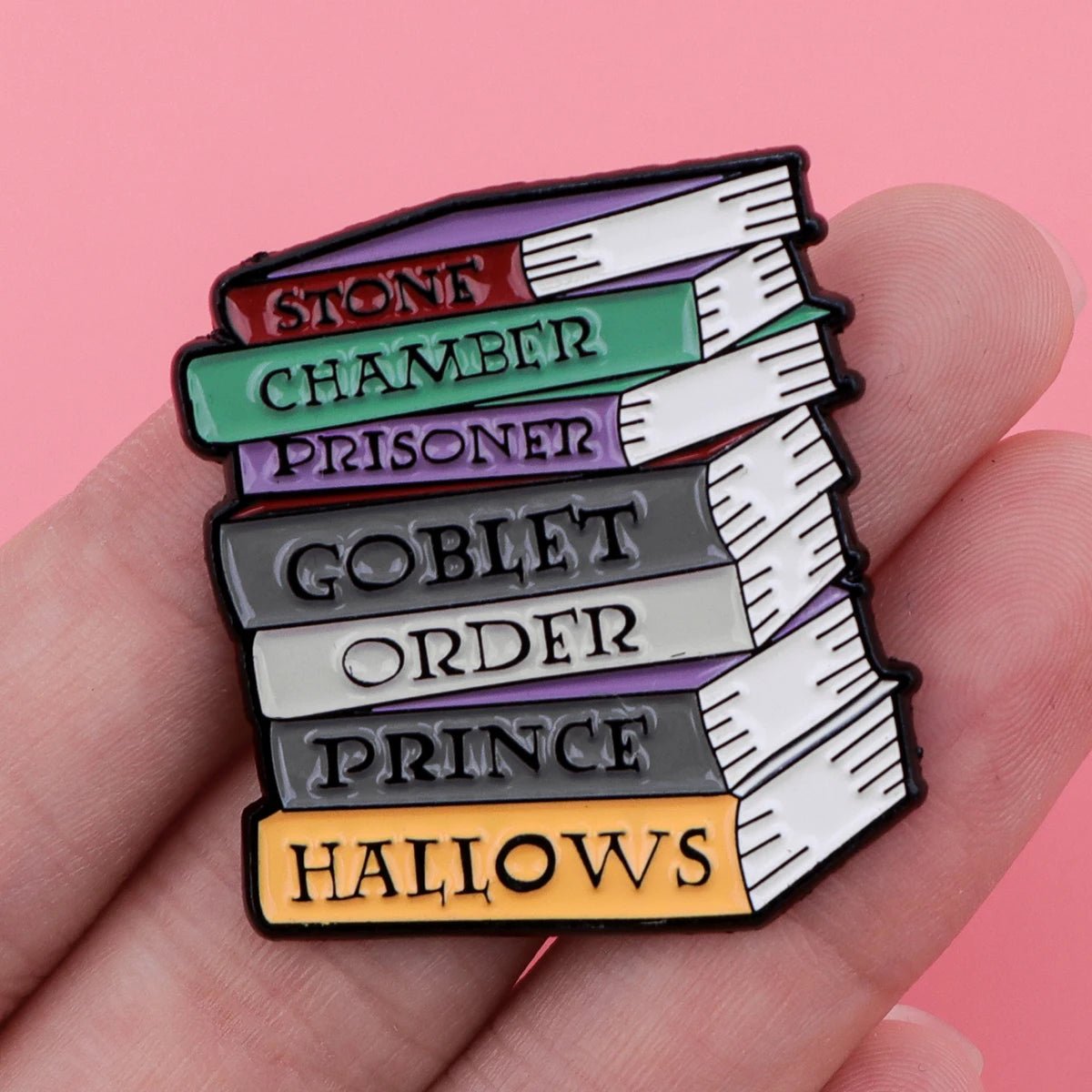 A hand holds an enamel pin shaped like a stack of books, each spine labeled with Harry Potter titles—perfect for any Booktrovert and fan of fictional characters. The background is pink.