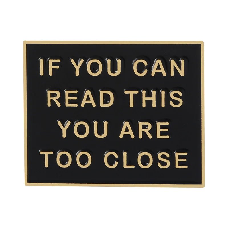 A black rectangular enamel pin by with gold trim and raised gold letters reading: IF YOU CAN READ THIS YOU ARE TOO CLOSE—a humorous statement accessory.