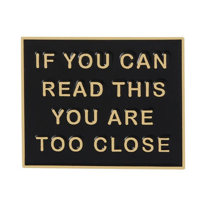 A black rectangular enamel pin by with gold trim and raised gold letters reading: IF YOU CAN READ THIS YOU ARE TOO CLOSE—a humorous statement accessory.