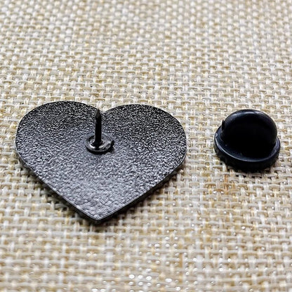 A black heart-shaped enamel pin by Kiss Me Later lies face down on woven beige fabric, displaying its pin and a detached black rubber back—an ideal addition for any enamel pin collector.