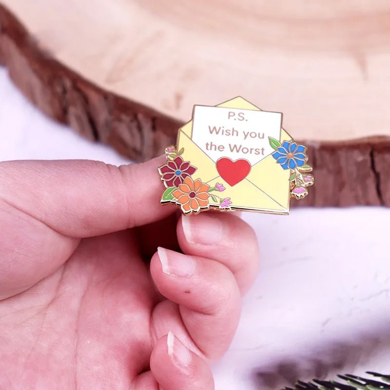 A hand holding yellow floral enamel pin, shaped like an envelope with flowers and a heart, featuring the phrase “P.S. Wish you the Worst.”.