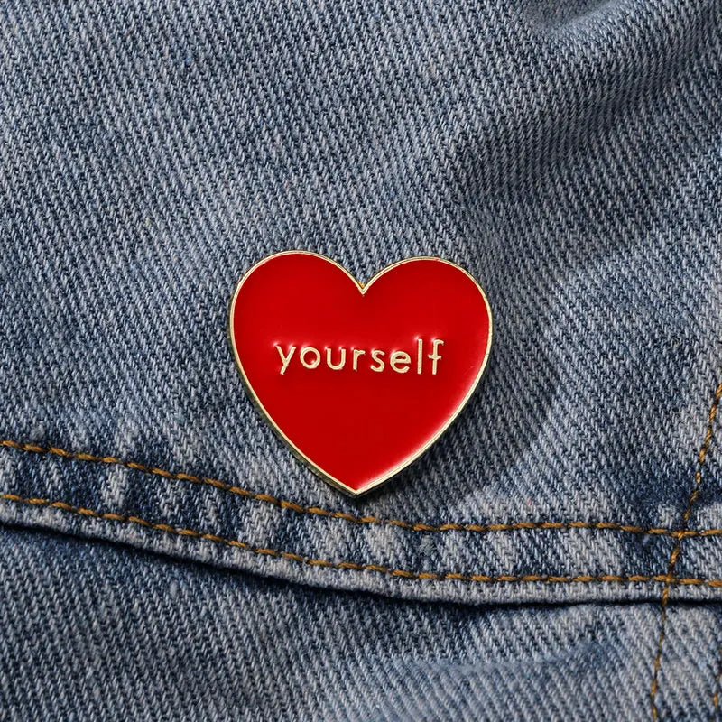 A red love heart enamel pin by Kiss Me Later, featuring the word “yourself” in lowercase white letters, is attached to blue denim fabric with visible stitching.