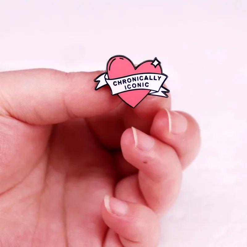 A hand holds a pink love heart enamel pin with a white banner that reads CHRONICALLY ICONIC, set against a pale pink background.