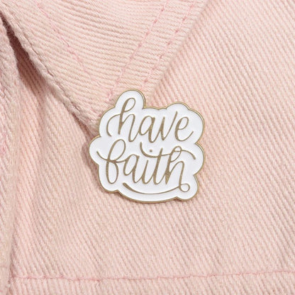 A white enamel pin with gold trim  featuring have faith in cursive, is attached to textured light pink fabric—a subtle, Bible-inspired reminder.