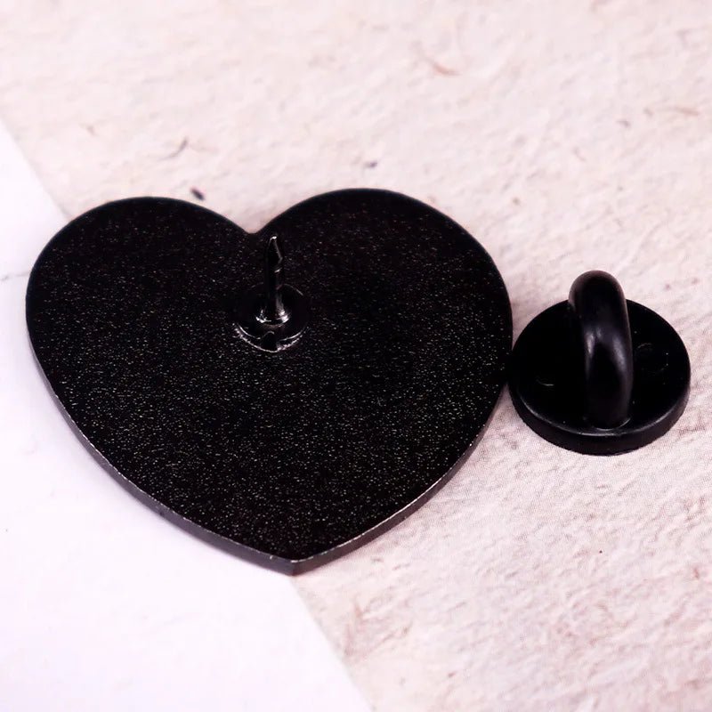A close-up of a Kiss Me Later black heart pin on a light textured surface, with its round backing set beside it.