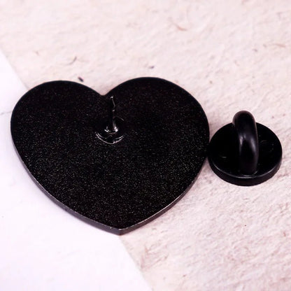 A close-up of a Kiss Me Later black heart pin on a light textured surface, with its round backing set beside it.