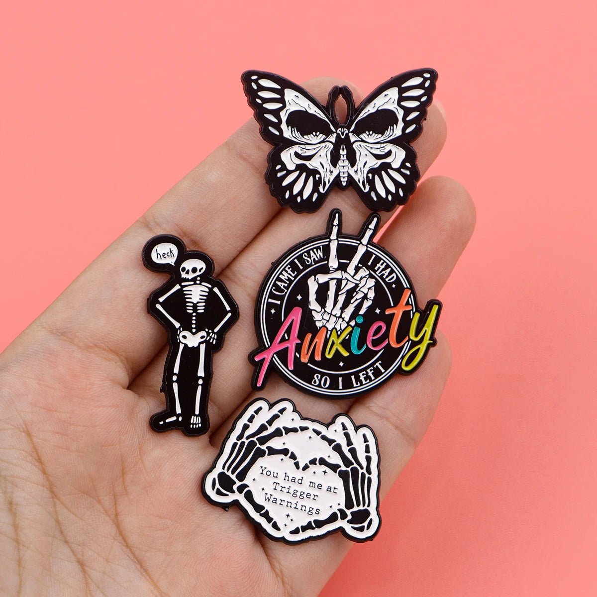 A hand holds four Kiss Me Later enamel pins: a black and white butterfly, a skeleton hand saying heh!, a peace sign reading “Anxiety so I left,” and two hands with You had me at trigger warnings on pink.