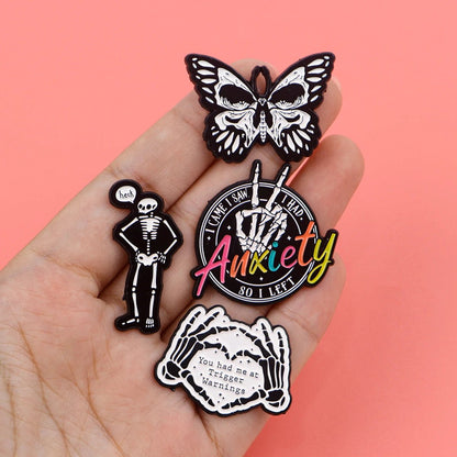 A hand holds four Kiss Me Later enamel pins: a black and white butterfly, a skeleton hand saying heh!, a peace sign reading “Anxiety so I left,” and two hands with You had me at trigger warnings on pink.