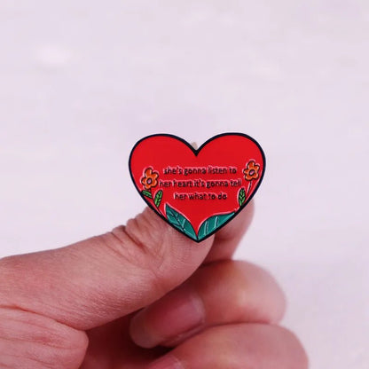 A hand holds an enamel pin shaped like a red heart with floral accents, featuring the text: shes gonna listen to her heart, its gonna tell her what to do.