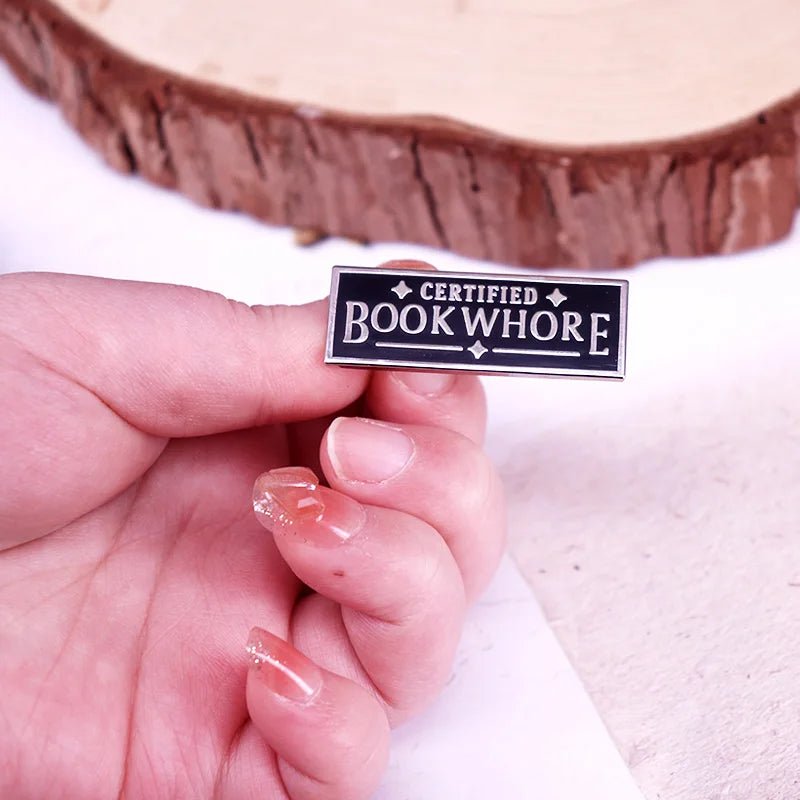 A hand with manicured nails holds a My Cutesy Era enamel pin that reads CERTIFIED BOOKWHORE in bold white letters on a black background.
