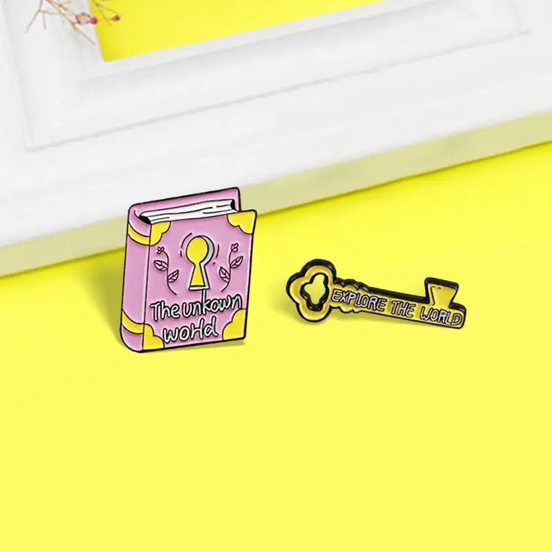 Enamel pins rest on a yellow surface: a pink The Unknown World book badge with a keyhole, and a yellow key pin that reads Explore the World.