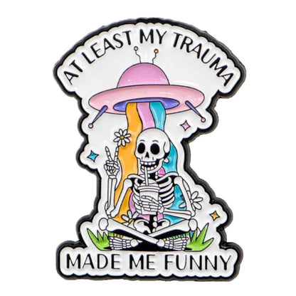 Enamel pin features a colorful skeleton sitting on grass, holding a drink and making a peace sign, with a rainbow pouring from a pink UFO overhead. The pin reads: AT LEAST MY TRAUMA MADE ME FUNNY.