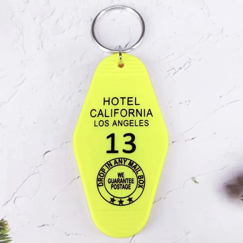 A yellow retro hotel key ring by Kiss Me Later, shaped like an old motel tag, reads HOTEL CALIFORNIA LOS ANGELES 13 with a circular stamp that says DROP IN ANY MAIL BOX - WE GUARANTEE POSTAGE.