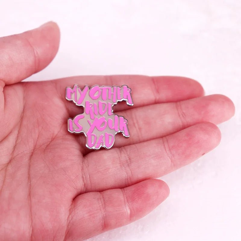 A hand holding a hot pink enamel pin with the words MY OTHER RIDE IS YOUR DAD in bold, playful letters—the perfect funny accessory for pin lovers.