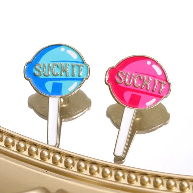 Two My Cutesy Era Suck It lollipop enamel pins, one blue and one pink, are displayed near a gold frame. Add playful flair to your collection with these unique pins from My Cutesy Era.