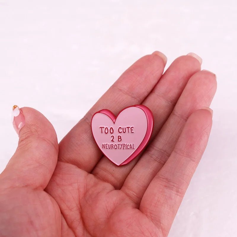A hand holds a pink candy heart pin by My Cutesy Era, featuring TOO CUTE 2 B NEUROTYPICAL in bold letters. The soft background highlights the sweet and playful neurodiversity design of this standout accessory.