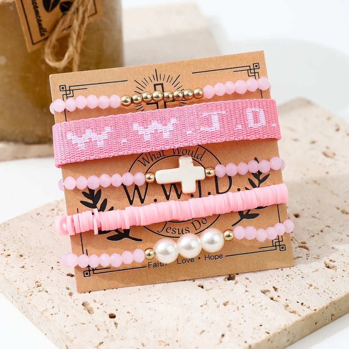 A pink bracelet stack by Kiss Me Later features four beaded bracelets on a “What Would Jesus Do?” card: one woven WWJD, one with a white cross, and two with mixed beads and pearls.