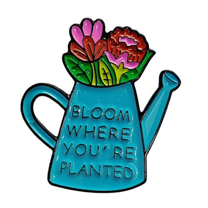 A My Cutesy Era enamel pin shaped like a blue watering can filled with pink and red flowers. The can features the phrase, Bloom where you’re planted, in newspaper-style text.