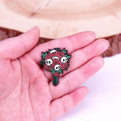 A hand holds an enamel pin shaped like a gothic bouquet of red roses, green leaves, and tiny white skulls, with a wooden surface in the background.