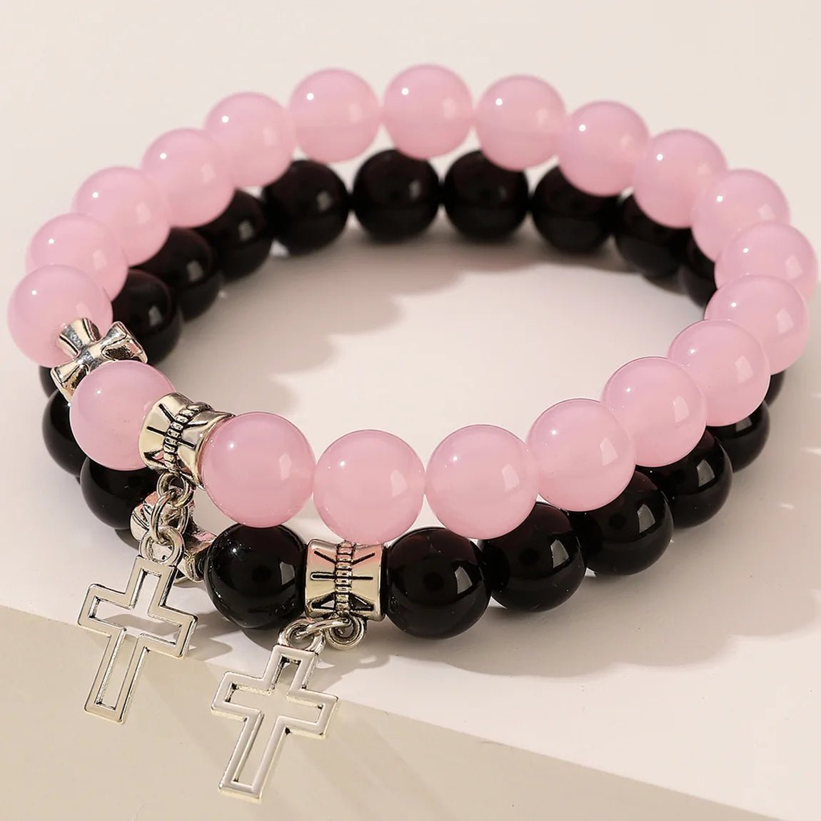 Kiss Me Later offers two crystal bead bracelets—one pink, one black—each with a silver cross charm, displayed on a white surface. Perfect Christian stone bracelets for any occasion.