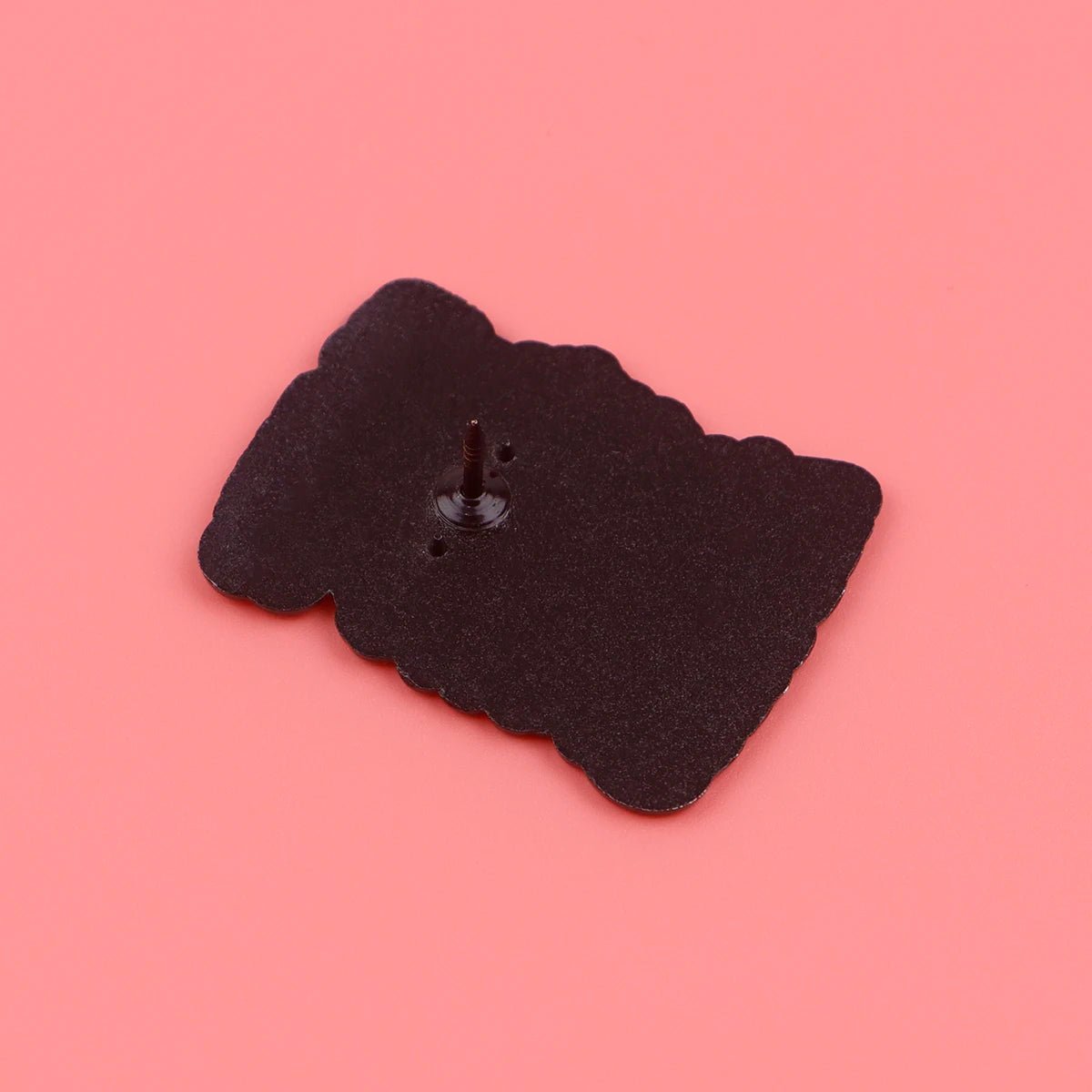 A Kiss Me Later black skeleton hand enamel pin with a single post and back clasp lies face down on a solid pink background, bringing subtle spooky vibes to your collection.