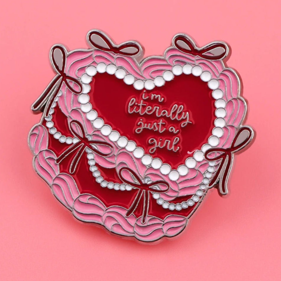 Enamel pin features a heart shape inspired by a pink love heart cake, with pink and red scalloped edges, bows, white pearl-like accents on a pink background, and Im literally just a girl in cursive.