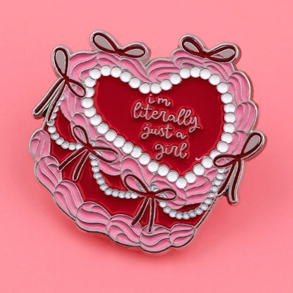 Enamel pin features a heart shape inspired by a pink love heart cake, with pink and red scalloped edges, bows, white pearl-like accents on a pink background, and Im literally just a girl in cursive.
