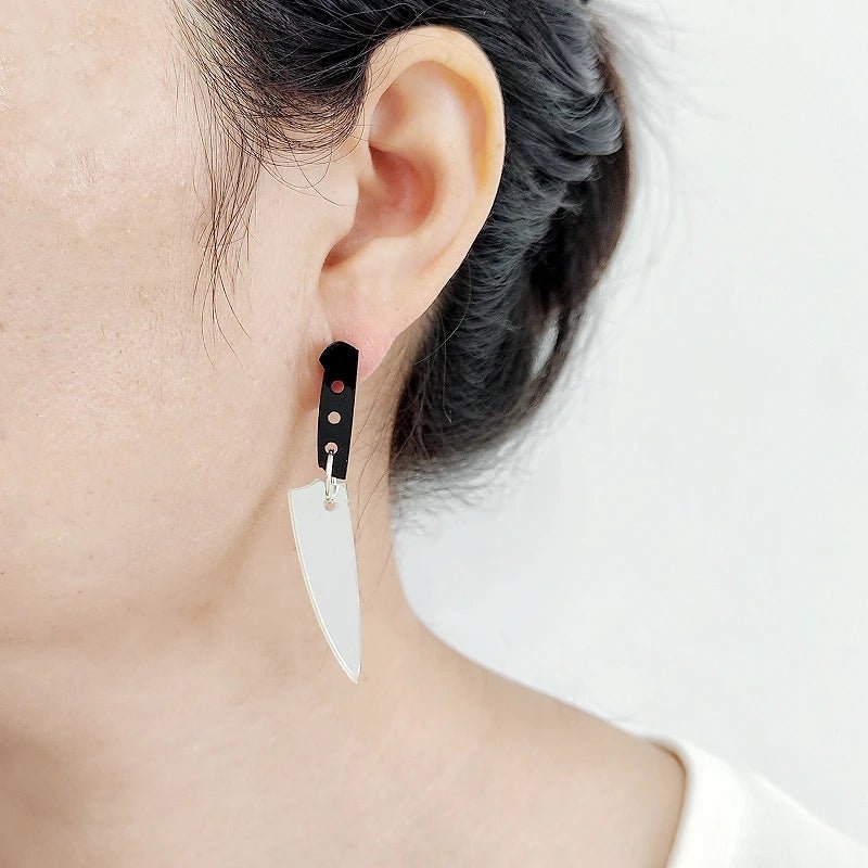 Close-up of a person with dark hair pulled back wearing Kiss Me Later knife earrings featuring acrylic charms—a silver blade and black handle—set against a plain white background.
