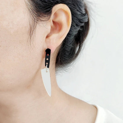 Close-up of a person with dark hair pulled back wearing Kiss Me Later knife earrings featuring acrylic charms—a silver blade and black handle—set against a plain white background.