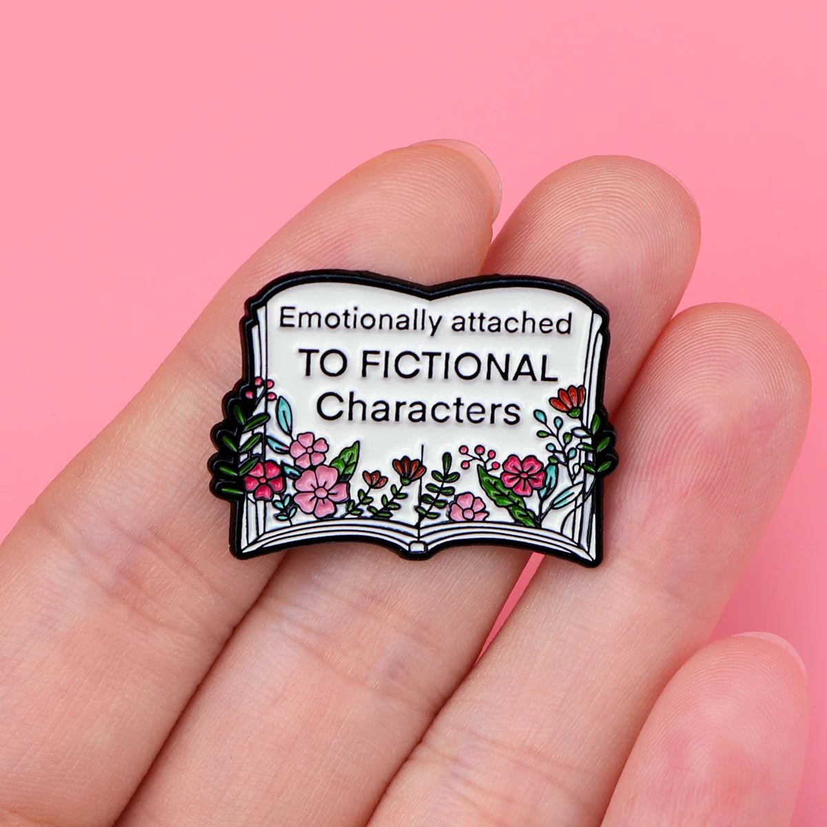A hand holds a pin shaped like an open book with floral decorations, reading Emotionally attached TO FICTIONAL Characters—a perfect accessory for every Booktrovert, shown against a pink background.