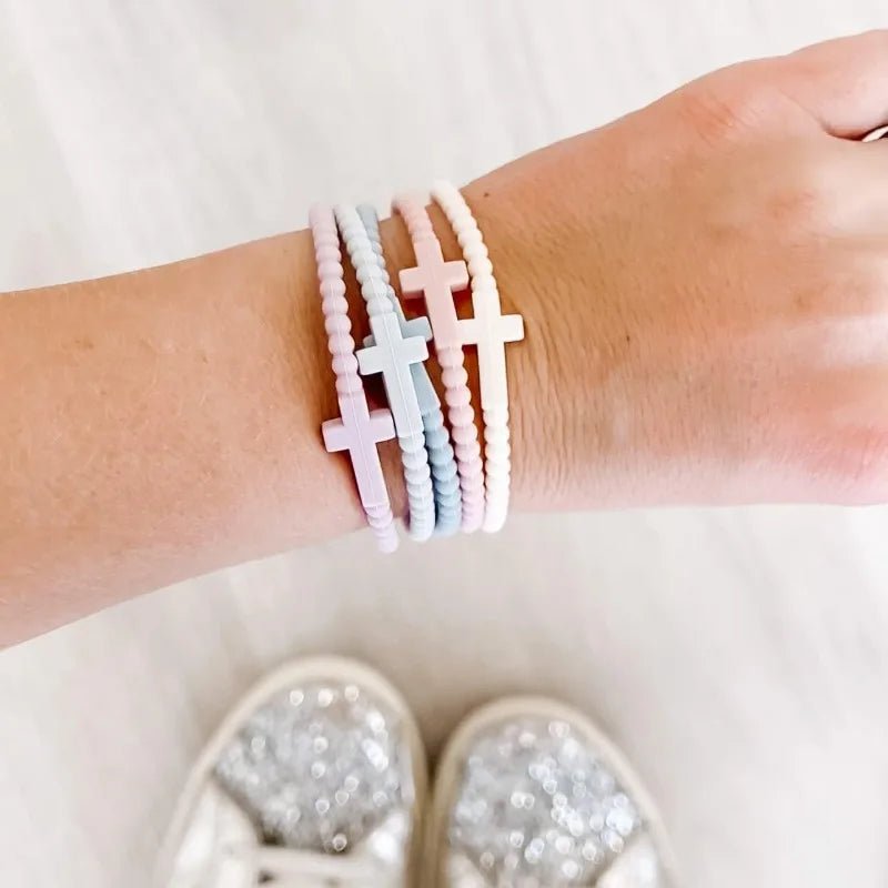 A hand shows off four pastel Kiss Me Later Cutie Bracelets with pink, blue, and white cross charms and silicone beads. Sparkly silver shoes are also worn.