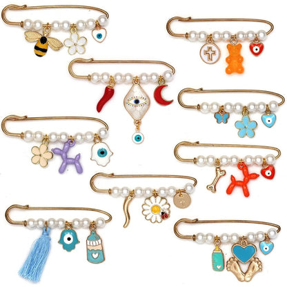 Eight gold safety pins by Kiss Me Later, adorned with pearls, beads, and whimsical brooch charms—flowers, animals, evil eye accents, hearts, stars, tassels—arranged on white. A charming choice for a baby shower gift.