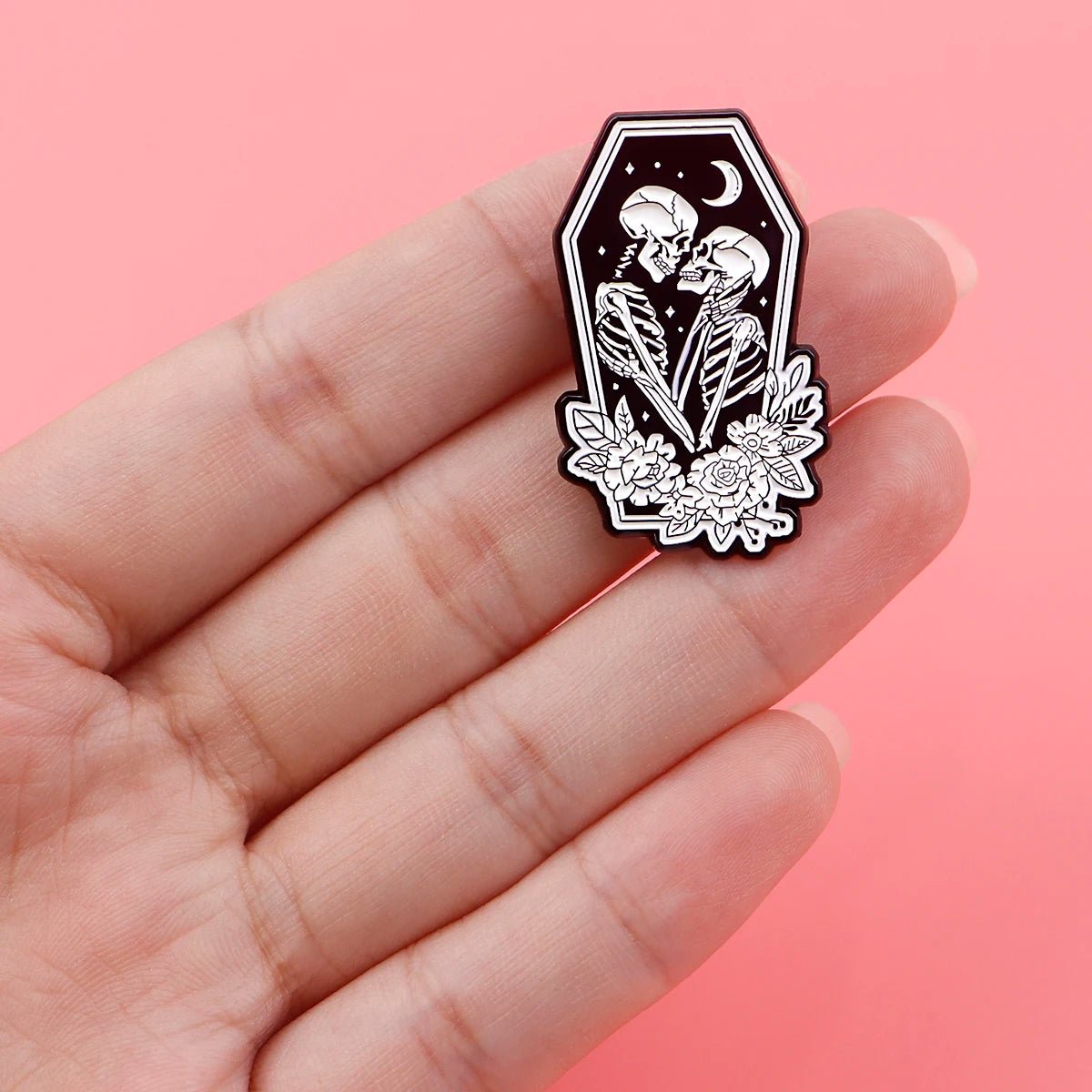 A hand displays the Kiss Me Later skeleton hand enamel pin, featuring two skeletons embracing and kissing inside a coffin with floral accents and a crescent moon, set against a pink background for spooky vibes.