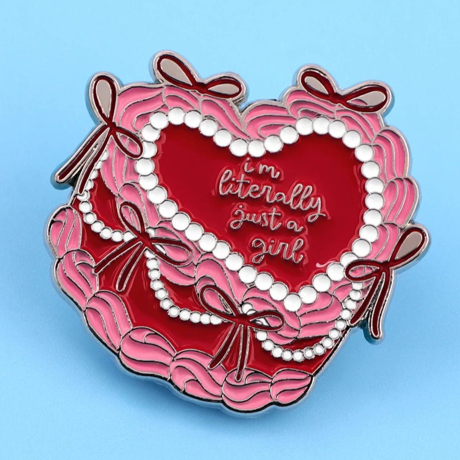 Heart-shaped cake pin features pink ruffles, pearls, and red bows with im literally just a girl in white script on a red background, displayed against a light blue surface.