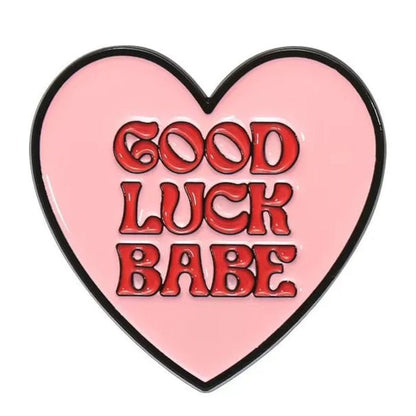 A pink heart enamel pin outlined in black features GOOD LUCK BABE in bold, red, bubbly letters at the center.