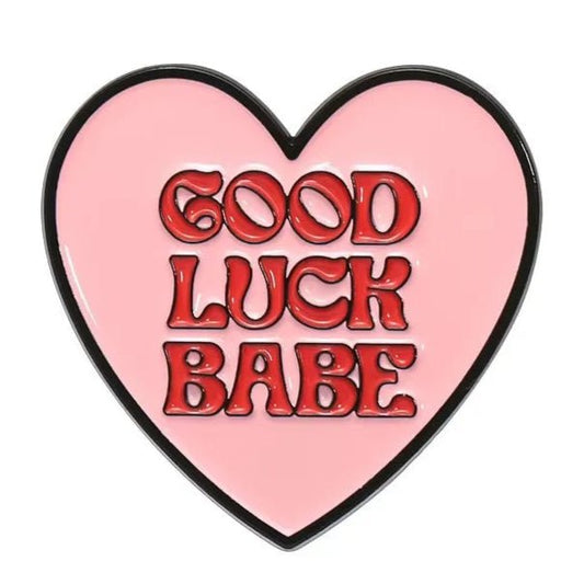 A pink heart enamel pin outlined in black features GOOD LUCK BABE in bold, red, bubbly letters at the center.