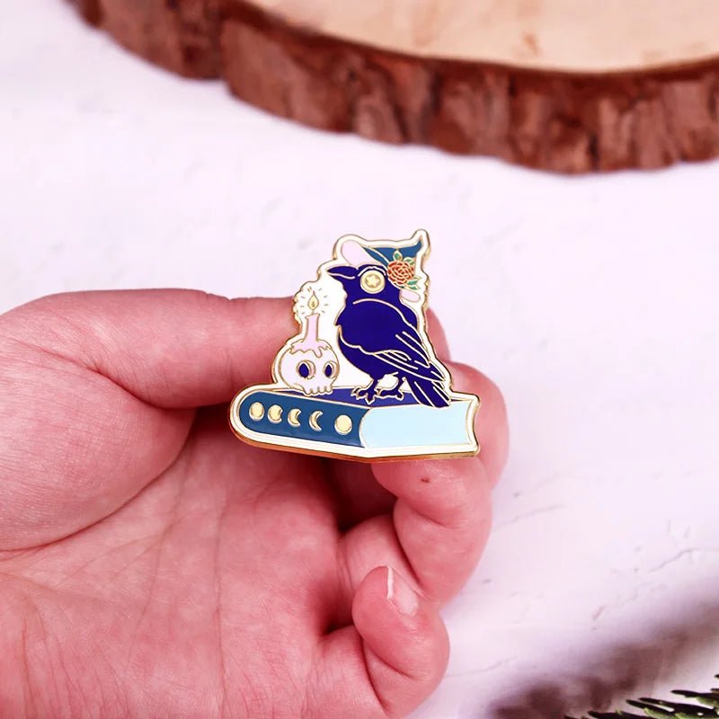 A hand displays an enamel pin of a dark blue raven on a closed book, with a white crystal ball, gold details, and a tiny candle accent, set against a light background.