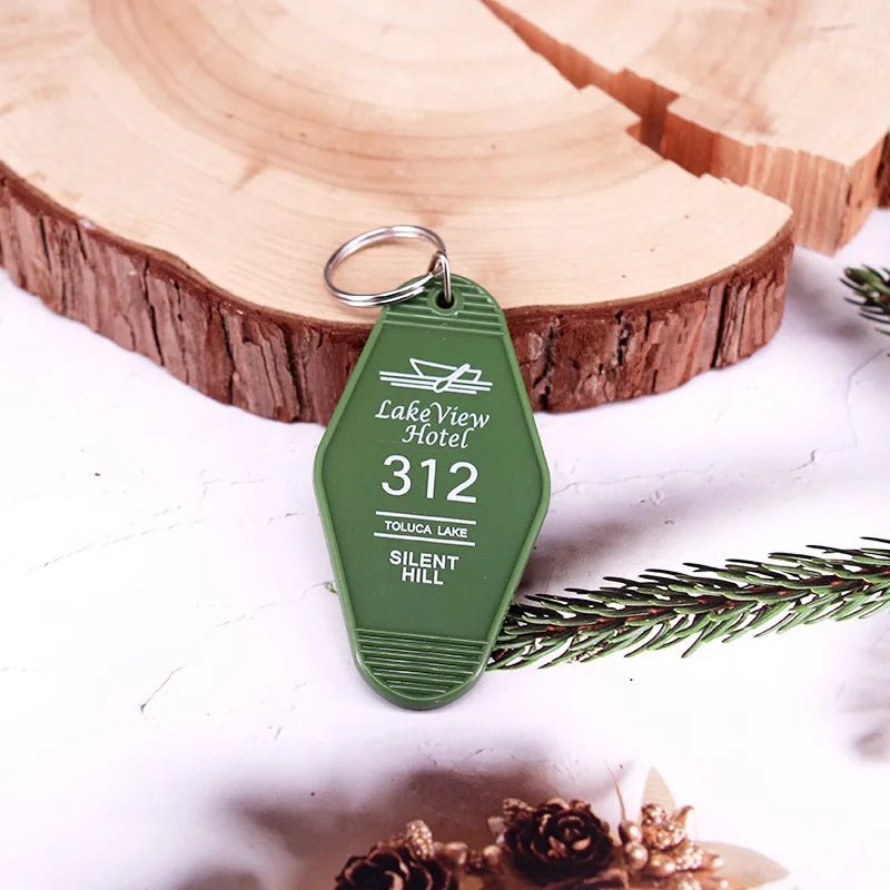 A green retro hotel key ring by Kiss Me Later, labeled LakeView Hotel 312, Toluca Lake, Silent Hill, rests on a white surface near a wooden slice and pine branch for a rustic, festive vibe.