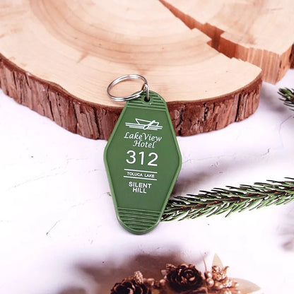 A green retro hotel key ring by Kiss Me Later, labeled LakeView Hotel 312, Toluca Lake, Silent Hill, rests on a white surface near a wooden slice and pine branch for a rustic, festive vibe.