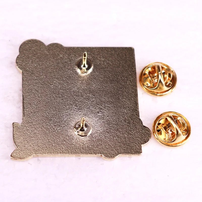 A square metallic literary enamel pin by Kiss Me Later, featuring two pointed back attachments and two gold-colored butterfly clutch pin backs, shown on a white surface.