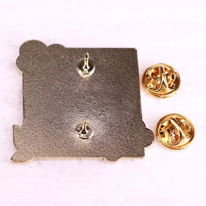 A square metallic literary enamel pin by Kiss Me Later, featuring two pointed back attachments and two gold-colored butterfly clutch pin backs, shown on a white surface.