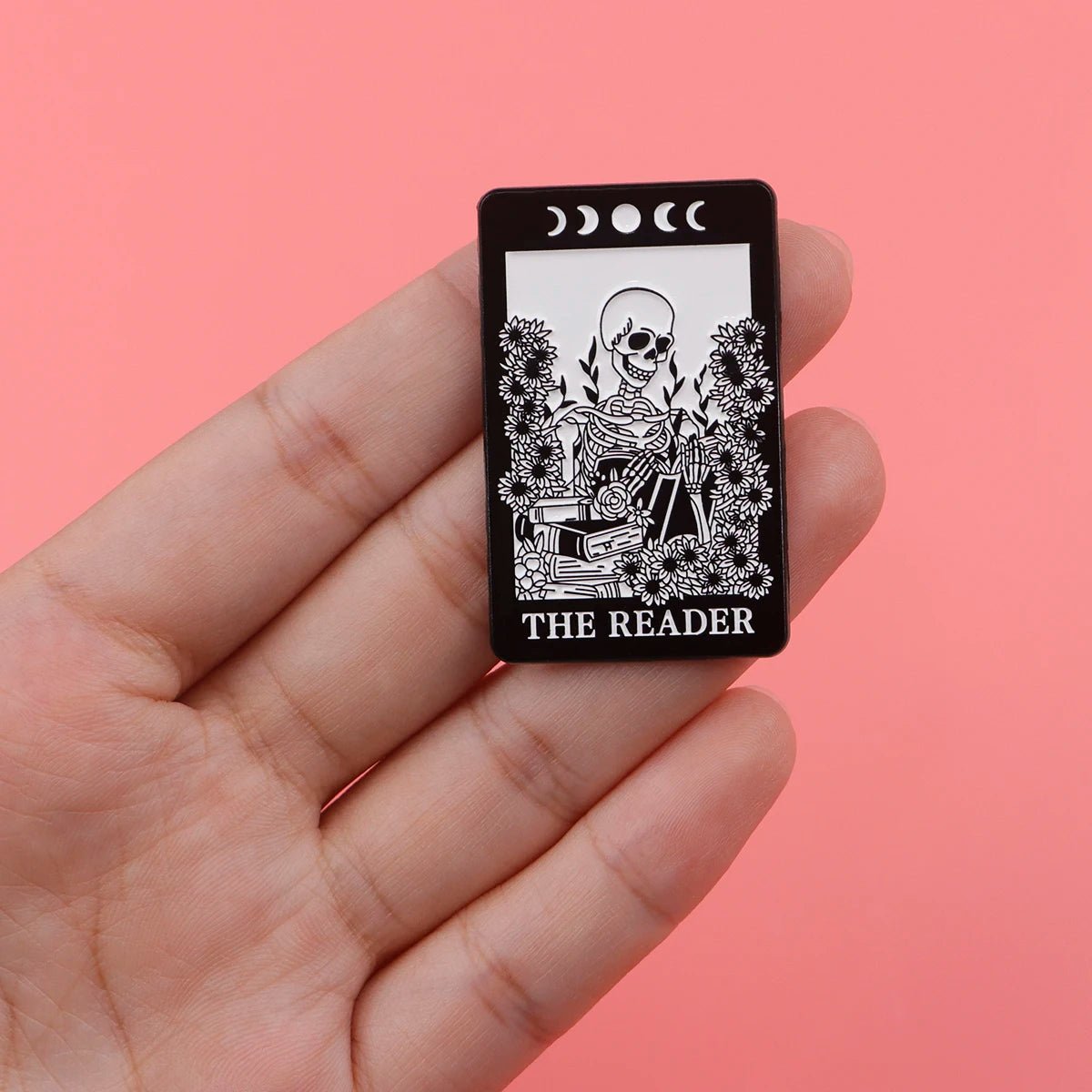 A hand holds a Kiss Me Later black enamel pin showing a skeleton reading among plants with moon phases above and THE READER at the bottom, set against a pink background.