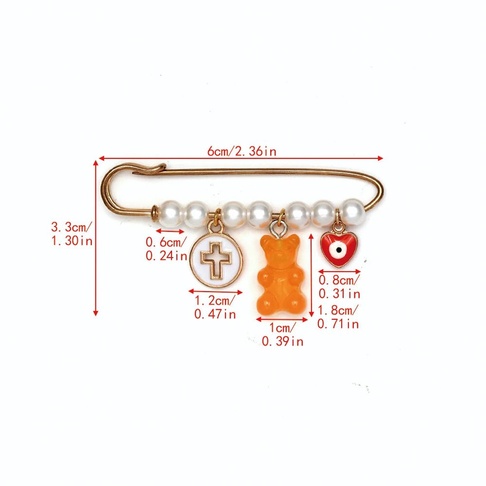 The Kiss Me Later gold safety pin brooch features five white beads and three dangling charms: a gold cross in a circle, an orange bear, and a red-and-white heart—an adorable baby shower gift. Dimensions in cm and inches included.